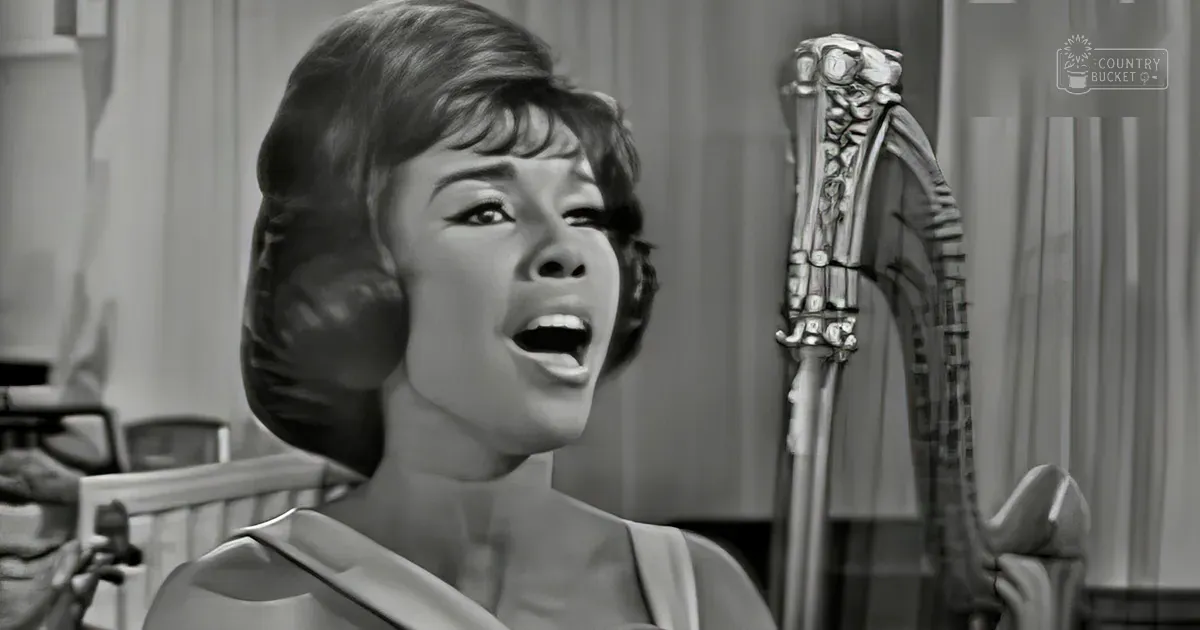 A young adult female in a vintage-style sleeveless top holding a microphone, possibly singing or speaking on stage with an ornate chair and curtains in the background.