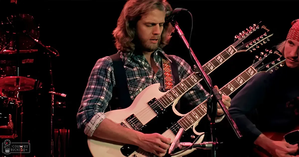 Live performance of a male musician in his young adult years wearing a plaid shirt and dark vest, playing a white electric guitar with another performer in the background.
