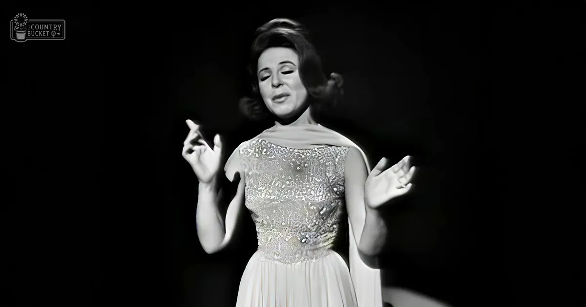 An adult female singer in an indoor setting, possibly a stage or studio, wearing a formal white dress with intricate embroidery and a fitted bodice and flared skirt, surrounded by minimalistic dark background lighting focused on her.
