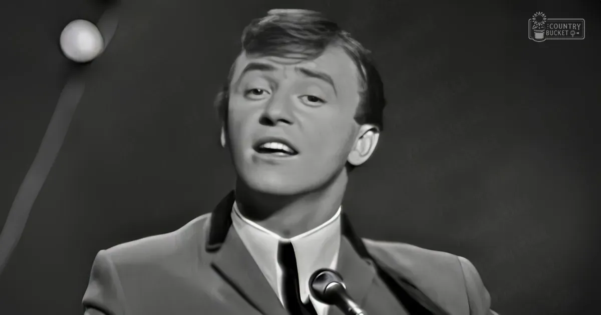 Young adult male singer wearing a dark formal suit with 'Madly Odd' text on his clothing, singing into a microphone in an indoor studio setting.