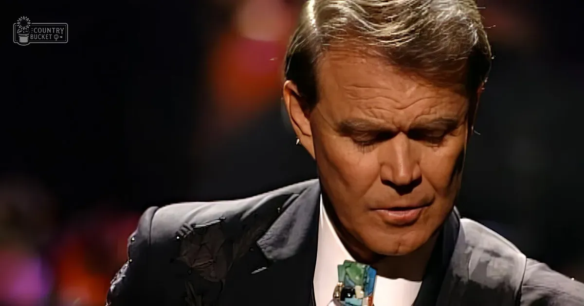 An older adult male in formal attire stands indoors under dim lighting with a blurred stage or audience area in the background.