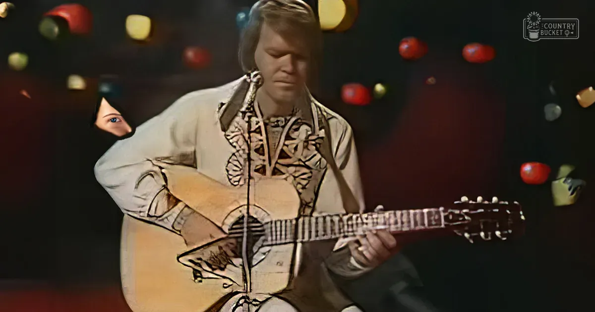 An adult male musician wearing a light-colored shirt with animal print patterns plays an acoustic guitar in a studio setting with dramatic lighting.