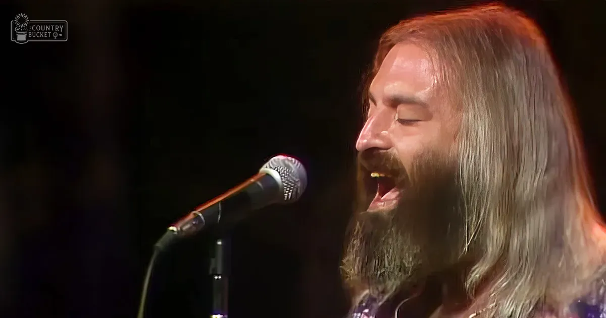 A middle-aged male singer wearing a patterned, low-cut garment in shades of purple and white at a music venue with stage lighting focused on him.