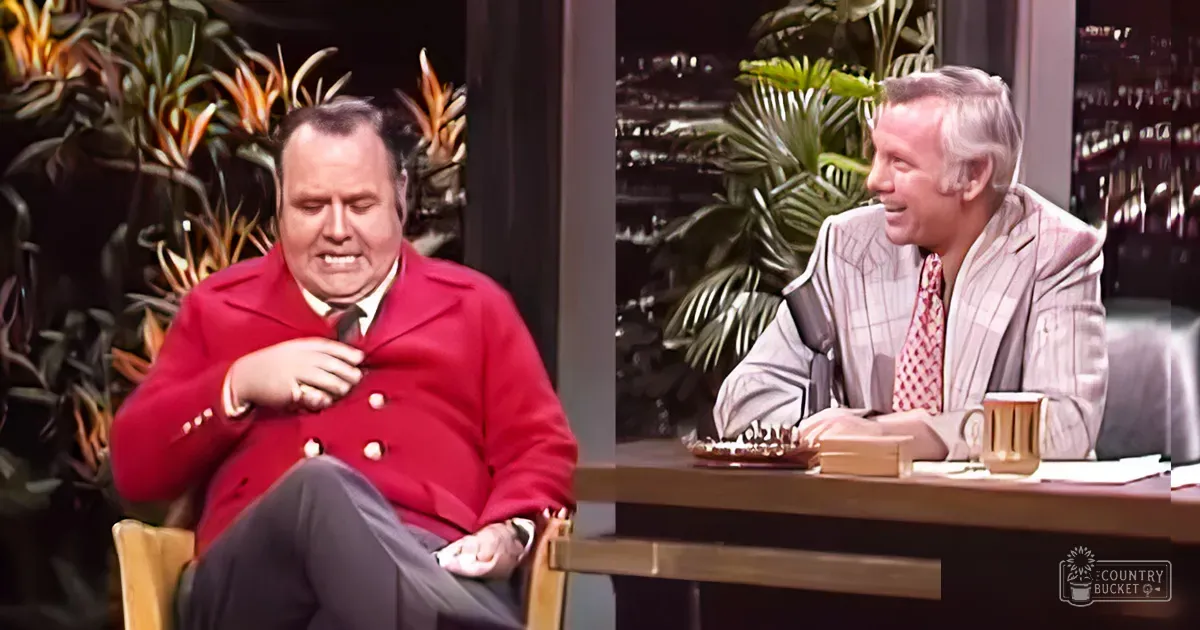 An indoor talk show set with a man in a red double-breasted coat and another older adult male in a beige pinstriped suit jacket. Wooden desk holds a coffee mug, papers, and game pieces against green plants.