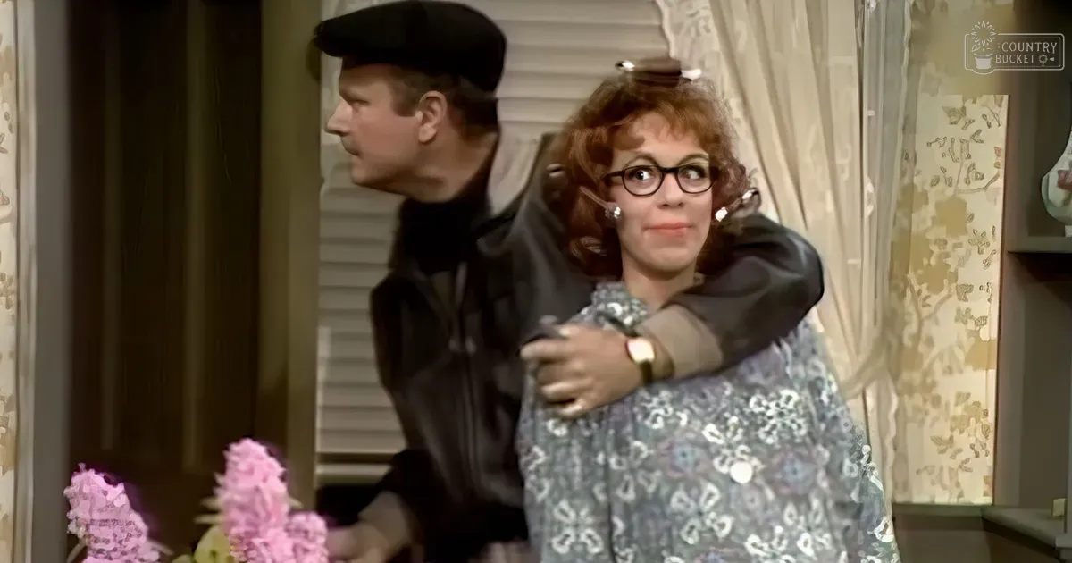 An adult male wearing a dark, possibly military-style jacket and black cap stands indoors with an adult female in a patterned blouse, black-rimmed glasses, and earrings. Floral wallpaper and lace curtains frame the scene.