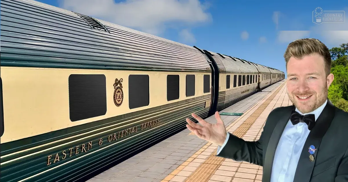 Adult male in a tuxedo with bow tie at an Eastern & Oriental Express train station platform surrounded by greenery on a bright, sunny day.