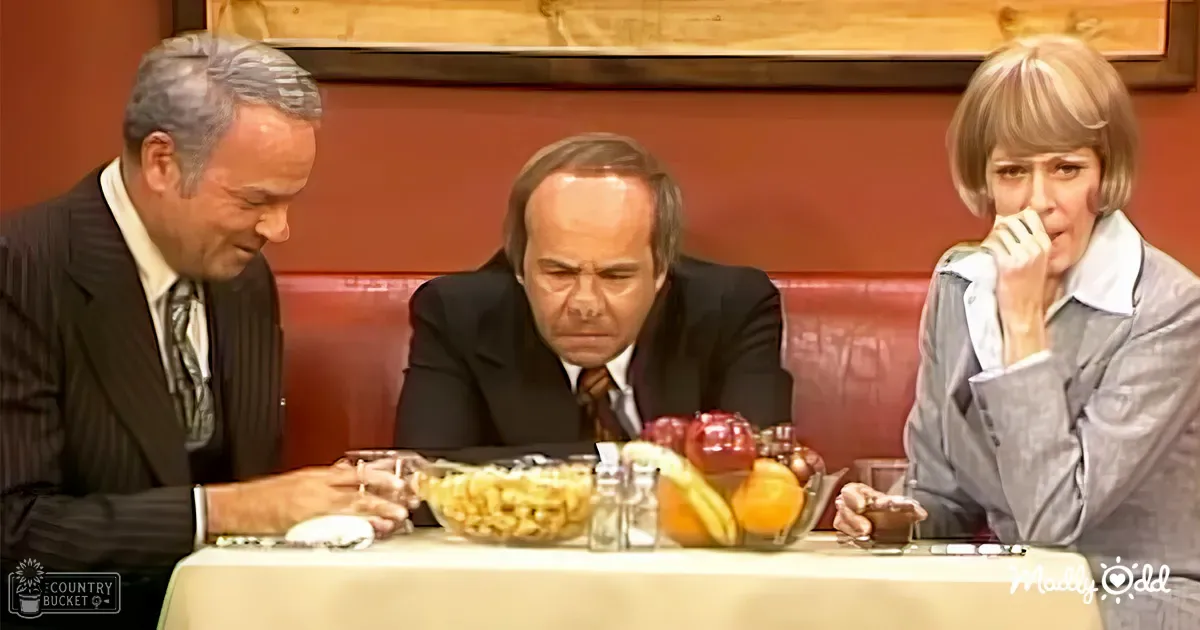 A middle-aged man in a black suit jacket and white dress shirt sits at a table with other diners, surrounded by mid-20th century-style restaurant decor.