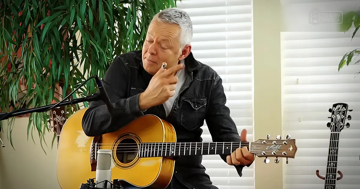 A middle-aged adult male wearing a black denim jacket playing an acoustic guitar in a home setting with another guitar, blinds, and potted plants.