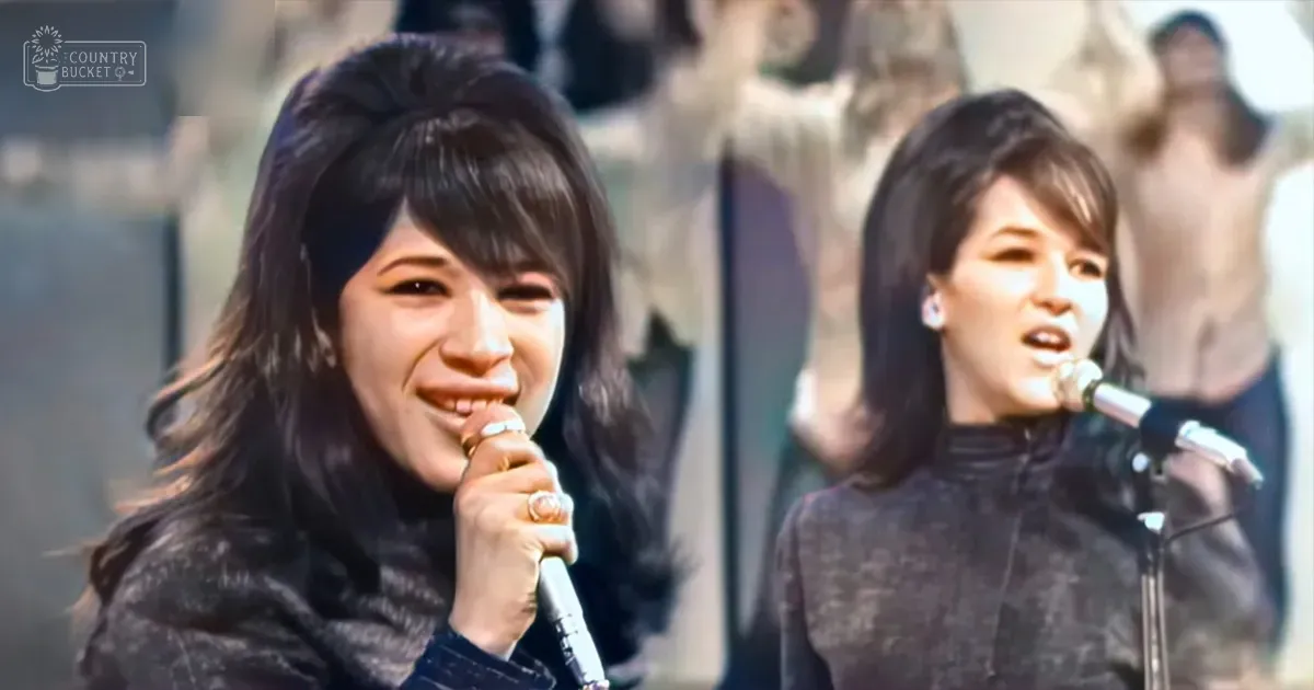 Two young adult female performers in dark textured jackets at an outdoor concert during daytime, wearing rings and surrounded by blurred figures and stone structures.