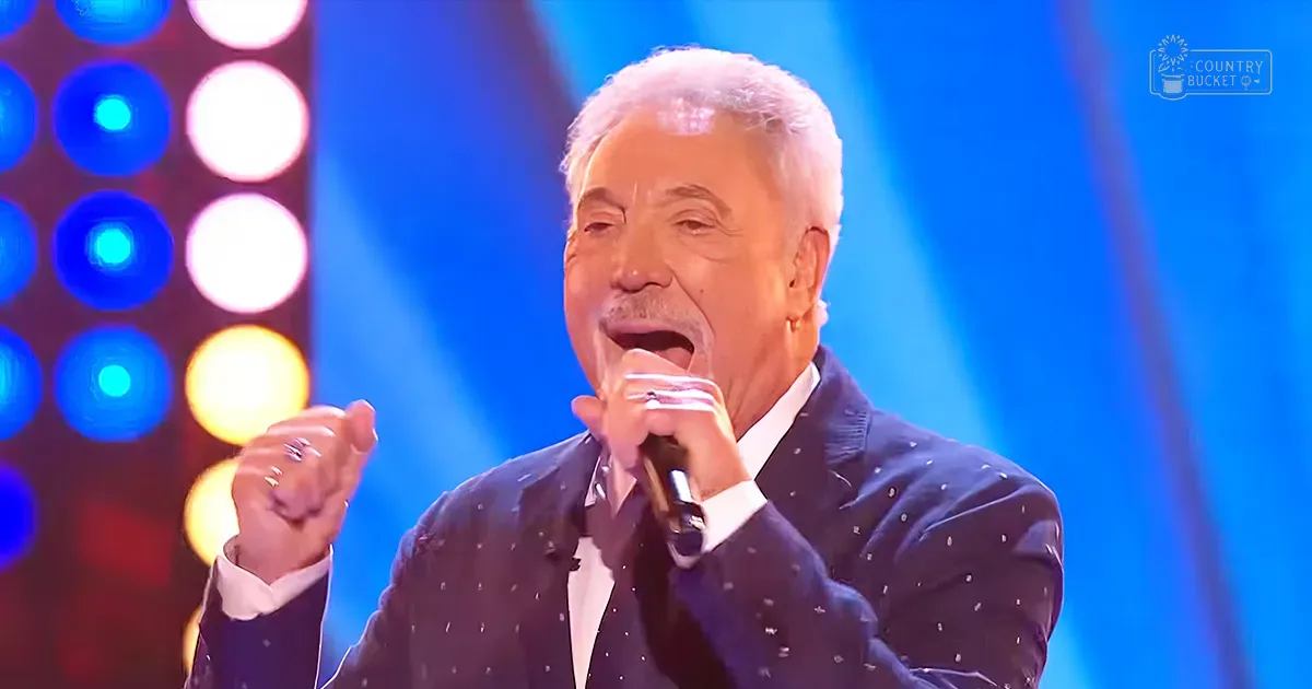 A middle-aged male singer in a dark blue suit with white polka dots, performing on stage at an event. He holds a microphone and wears a ring on his left hand's ring finger.