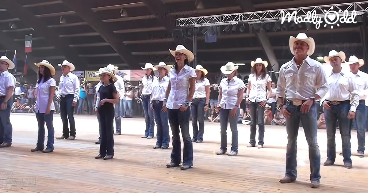 A group event at an indoor arena, possibly a rodeo, with various adults dressed in white shirts, blue jeans, cowboy boots, and hats, set against wooden flooring and metal beams under artificial lighting.