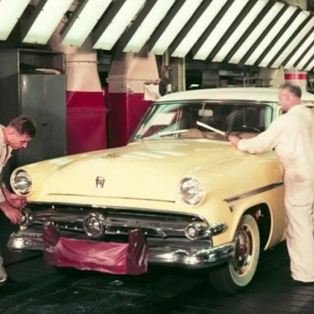 An older adult male in a white jumpsuit stands beside a vintage 1950s cream car in an indoor garage with striped awning and industrial equipment.