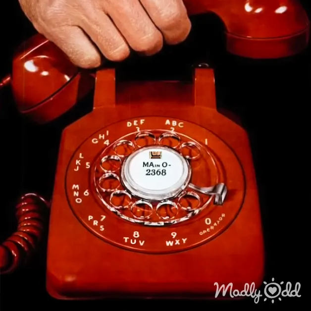 An old-fashioned red rotary telephone with a coiled cord, held by part of a hand indoors.