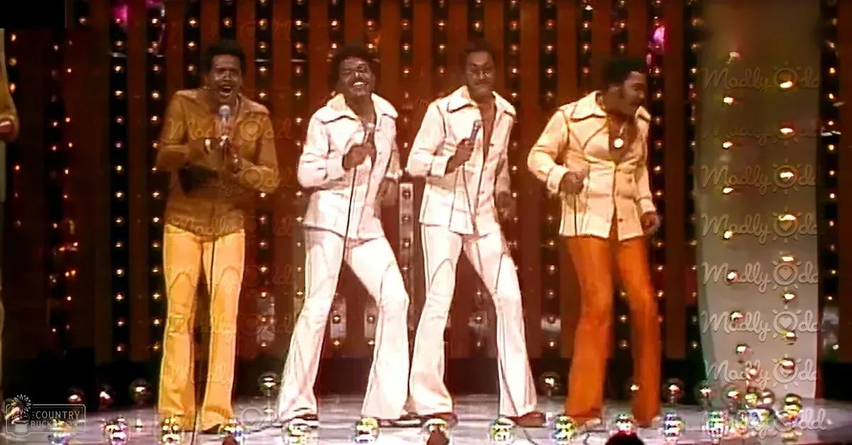 A musical performance group dressed in 1970s-style suits on a stage with vintage lighting and reflective spheres.