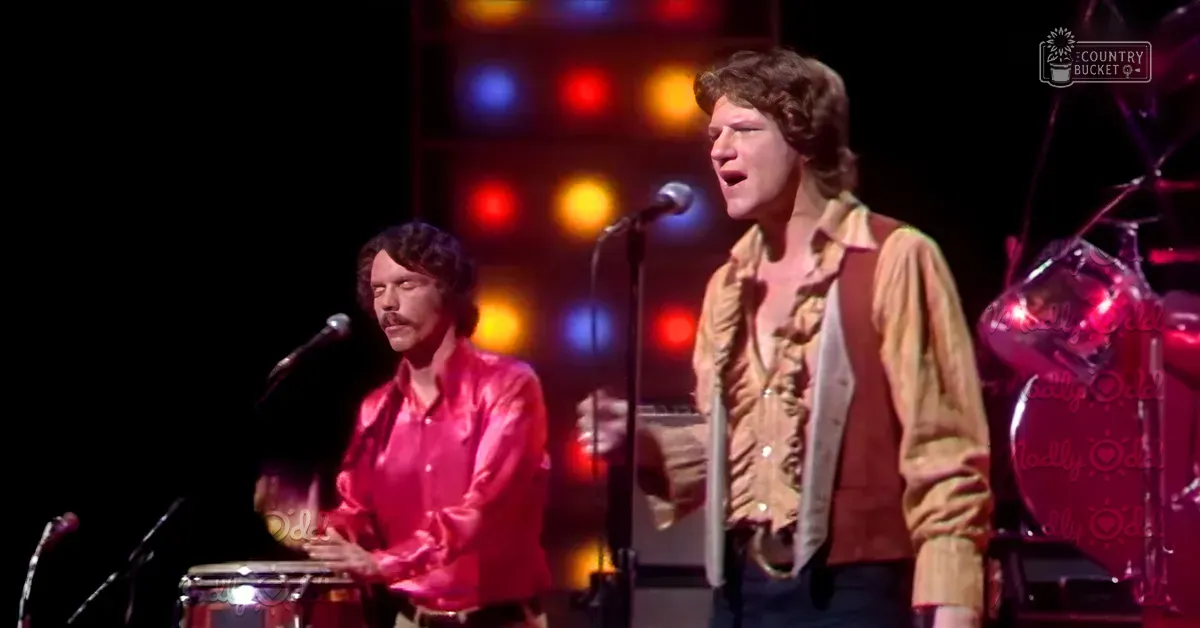Young adult male singers performing on an indoor stage with colorful lighting, wearing shiny pink button-up shirts and patterned yellow and brown shirts under vests.