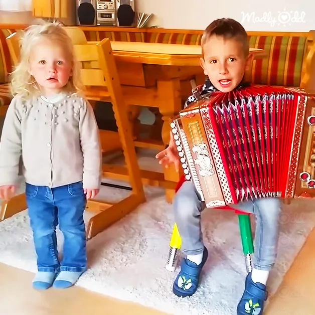 A toddler girl plays an accordion while a child boy looks on, set in a cozy home environment with wooden furniture and natural light.