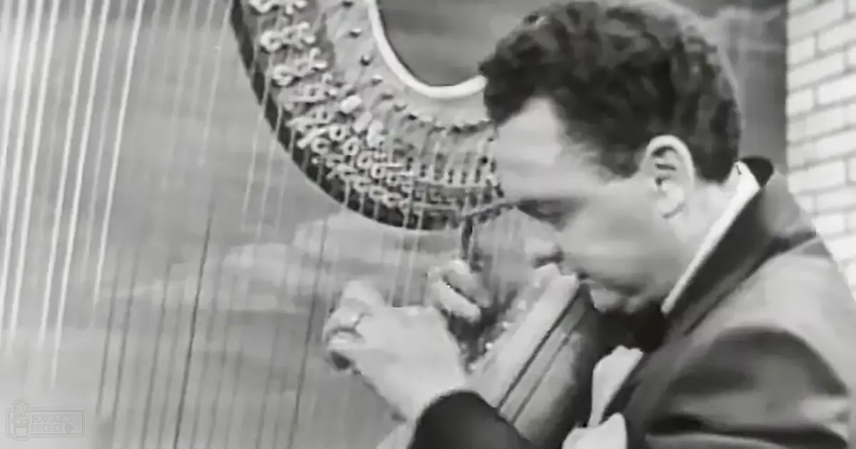 A young adult male playing a harp outdoors against a brick wall.