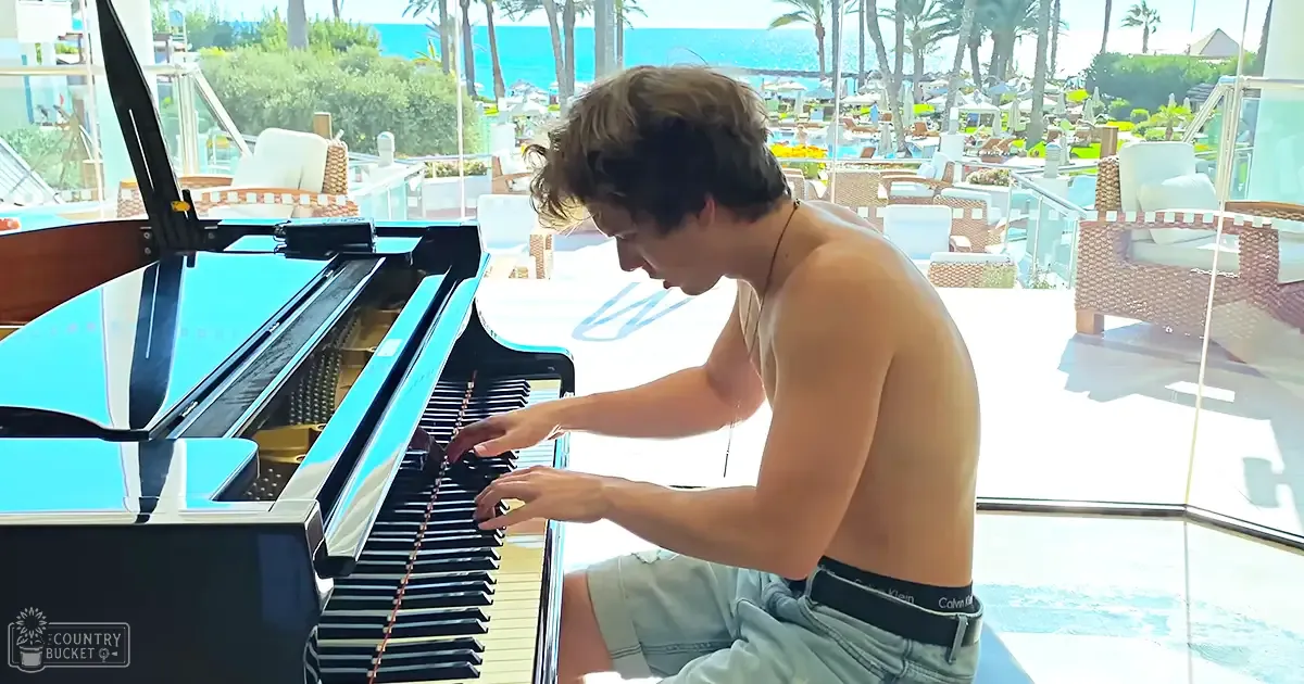 A young adult male plays a black grand piano on an outdoor patio overlooking the beach with palm trees and clear skies.