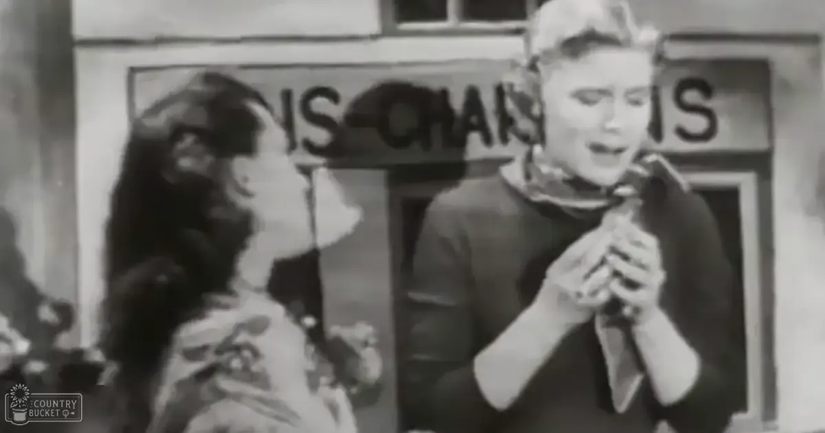 A black and white scene from an older film or television show set in a town square, with two adults dressed in mid-20th-century fashion under natural daylight.