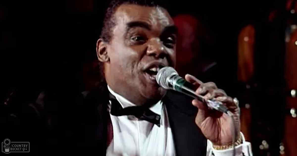 A middle-aged male singer in a black tuxedo with white shirt and bow tie performs into a microphone at an indoor venue likely during the evening or night.