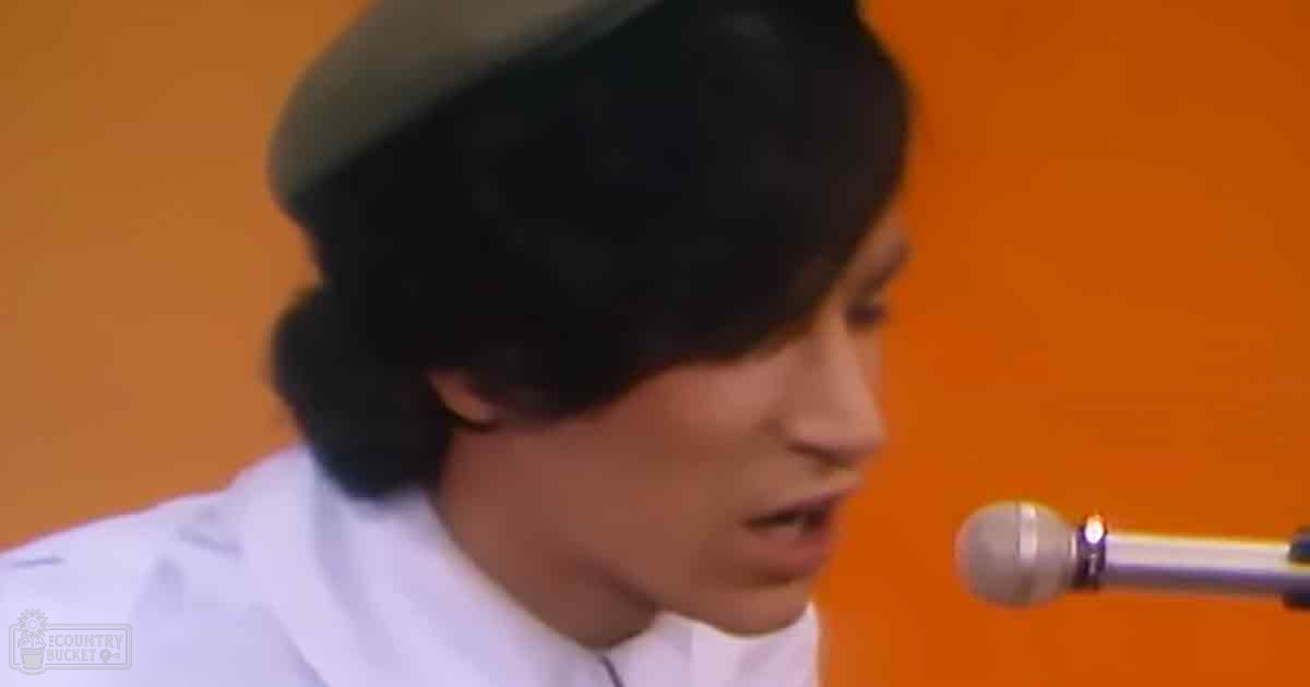 Young adult male wearing a relaxed fit, collared, long-sleeved white shirt with a dark-colored beret-style hat in an indoor setting with an orange background and a microphone for recording or broadcasting.