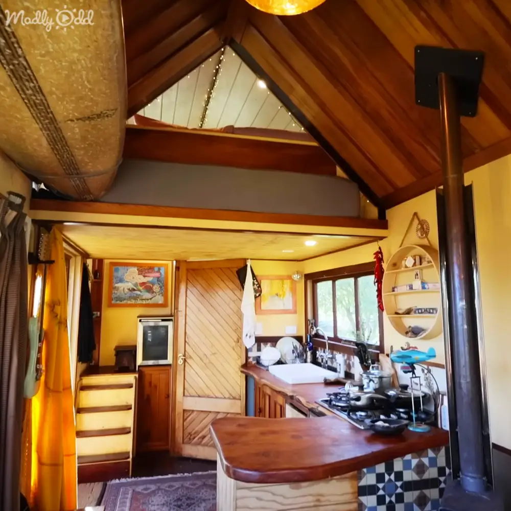 A cozy cabin interior with wooden furniture, a small refrigerator, gas stove, circular shelf, surfboard hanging from the ceiling, and a small rug on the floor near an entrance with natural light streaming through a window.