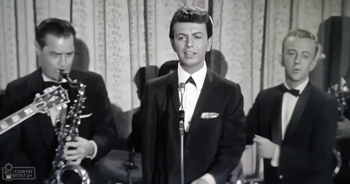 A young adult male singer wearing a dark suit jacket with a white shirt and black bow tie, holding a saxophone in an indoor studio setting.