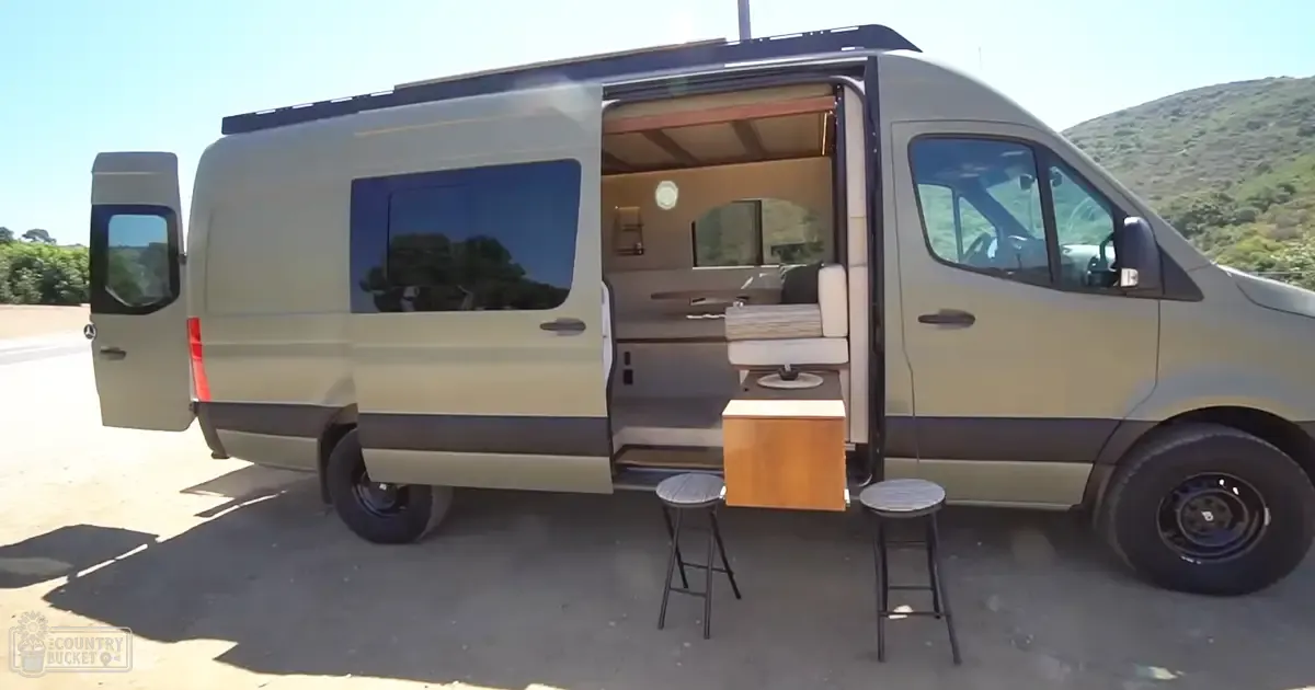 Beige Mercedes-Benz Sprinter van with open rear door, two black bar stools outside, compact living space interior visible, hills in natural outdoor setting, bright daylight