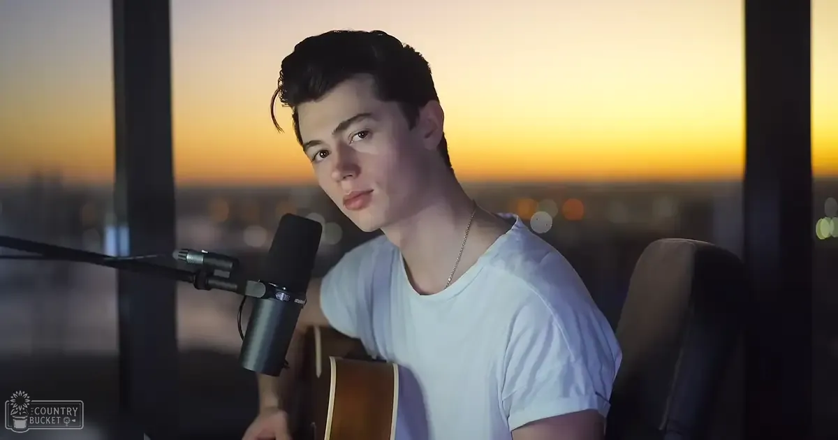 A young adult male wearing a white V-neck t-shirt, holding an acoustic guitar and a necklace, with a microphone in front of him, set against an urban cityscape background at dusk or dawn.