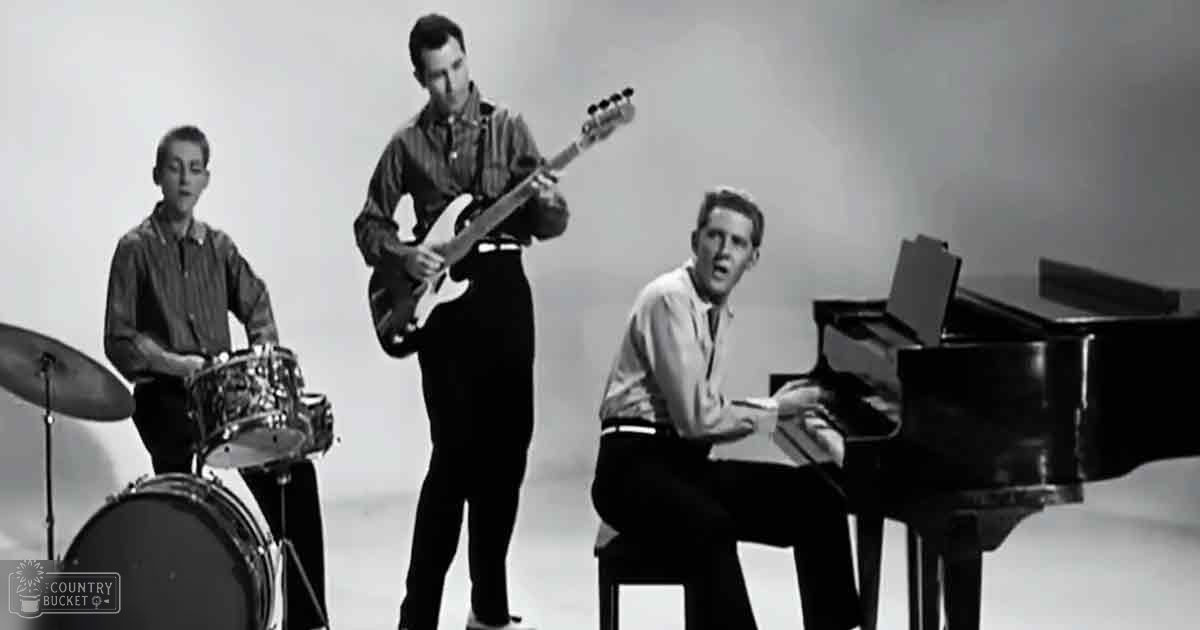 A musical performance scene with young adult males performing, surrounded by a drum set and an electric guitar, in a plain indoor studio setting.