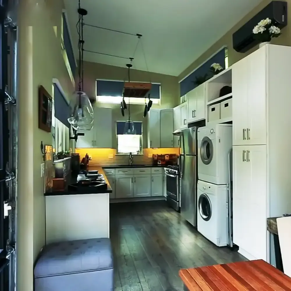 A residential home interior with white cabinets, a stainless steel refrigerator, an oven, a washing machine, dryer, wooden floor, pendant lights, and kitchen items on the counter.