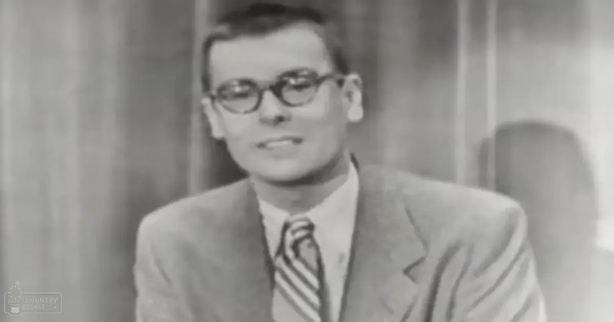 A young adult male wearing a tailored suit jacket with a collared shirt, long sleeves likely made of wool or silk, and a striped tie stands indoors by curtains.