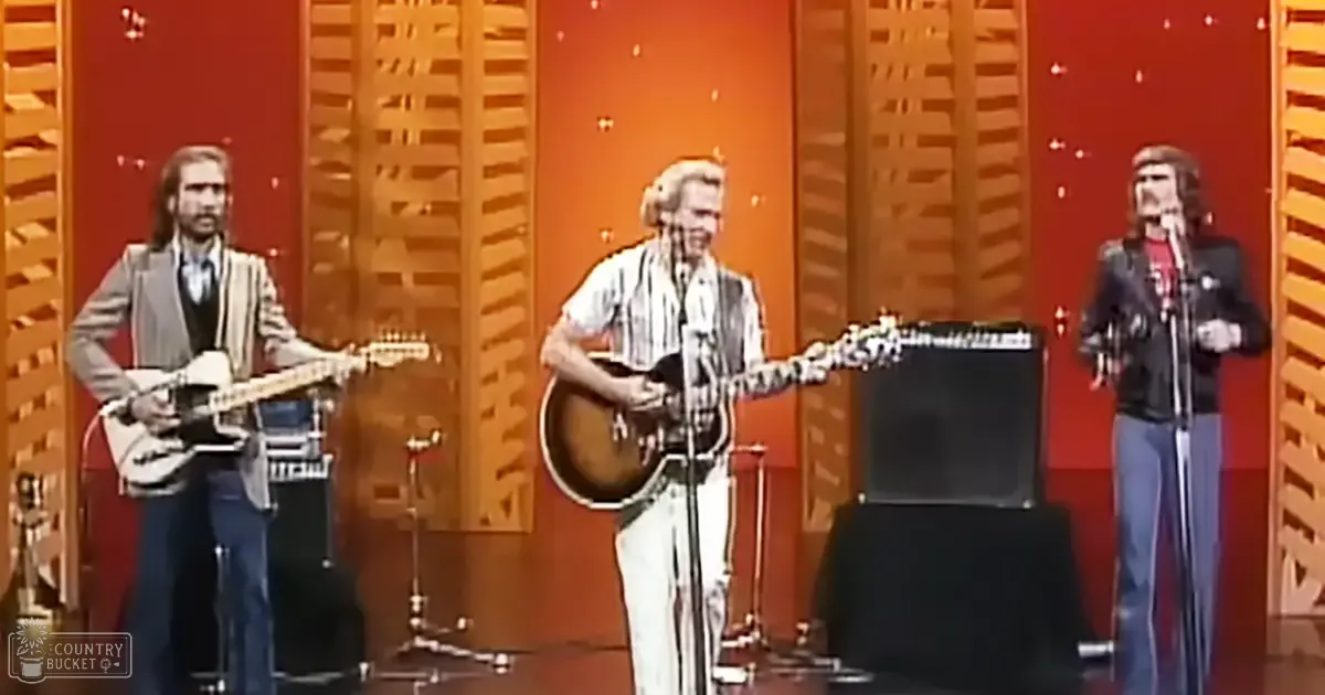 A musical performance from the 1970s with three adult male musicians on stage, playing electric guitars and singing into microphones. The setting is indoors, possibly a studio or theater with decorative wooden panels and red curtains.