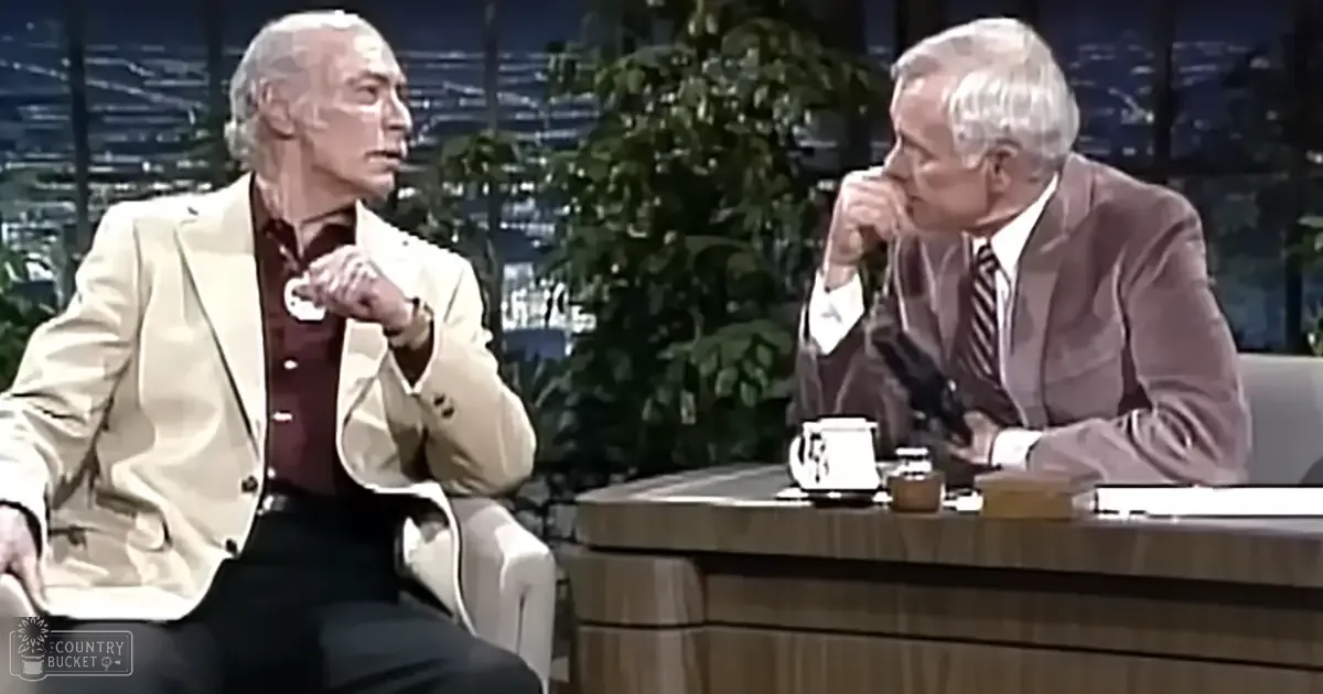 An older adult male talk show host wearing a light-colored blazer and an older adult guest in a dark suit jacket, seated at a wooden desk with books and plants visible in the background of a television studio setting.