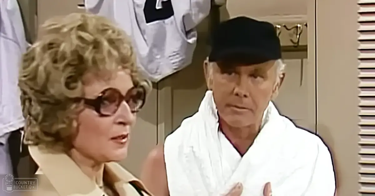 A middle-aged woman wearing a beige, vintage-style jacket with large round sunglasses stands indoors near a closet filled with garments and sports jerseys. The natural light suggests daytime.