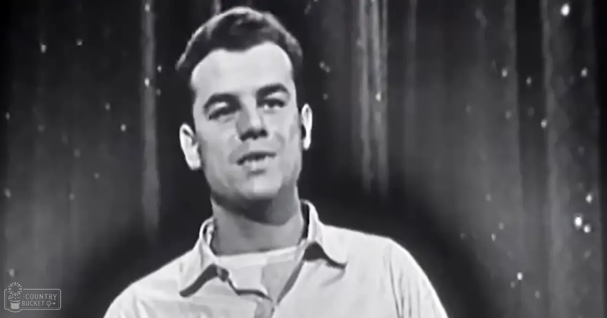 A young adult male in a relaxed, casual collared shirt with buttons and short sleeves, indoors against dark curtains or drapes.