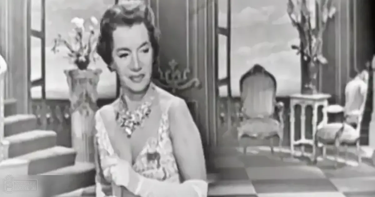 A woman wearing a loose-fitting floral blouse with ruffled sleeves and a statement necklace, seated indoors near furniture and home decor items.