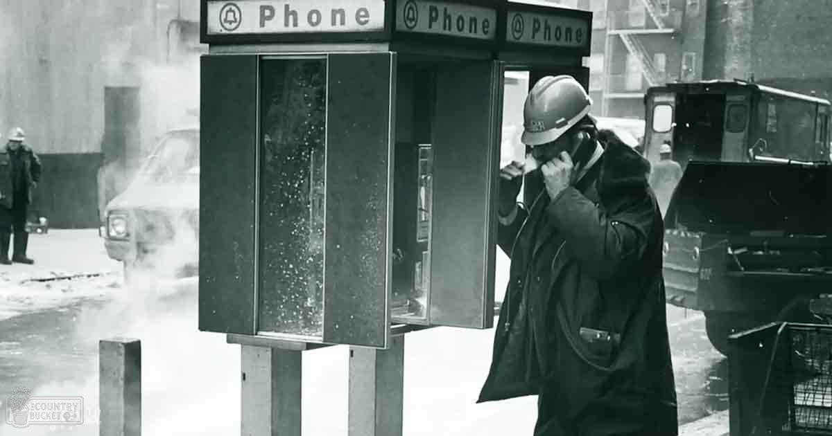 A man wearing dark coat and helmet in an urban winter setting using a payphone.