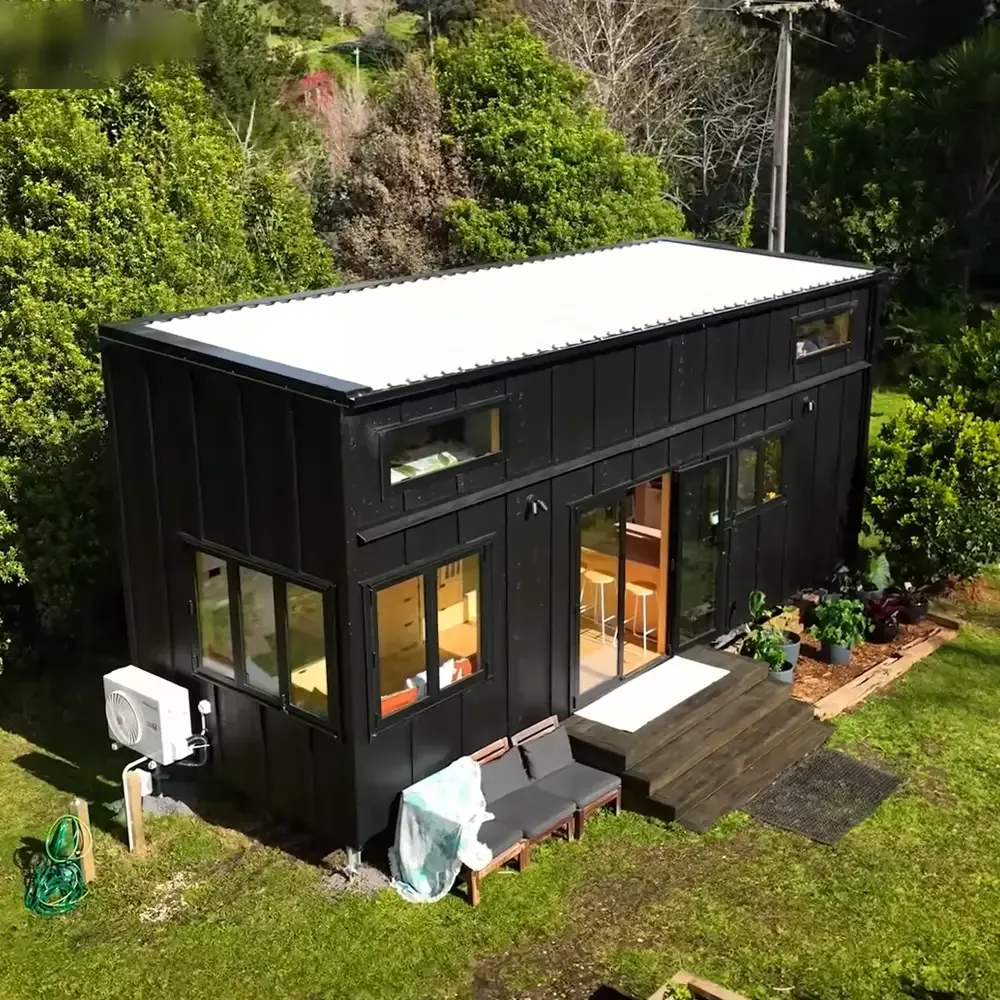 A modern tiny house nestled in a lush, green outdoor setting with potted plants and wooden furniture, suggesting spring or summer.