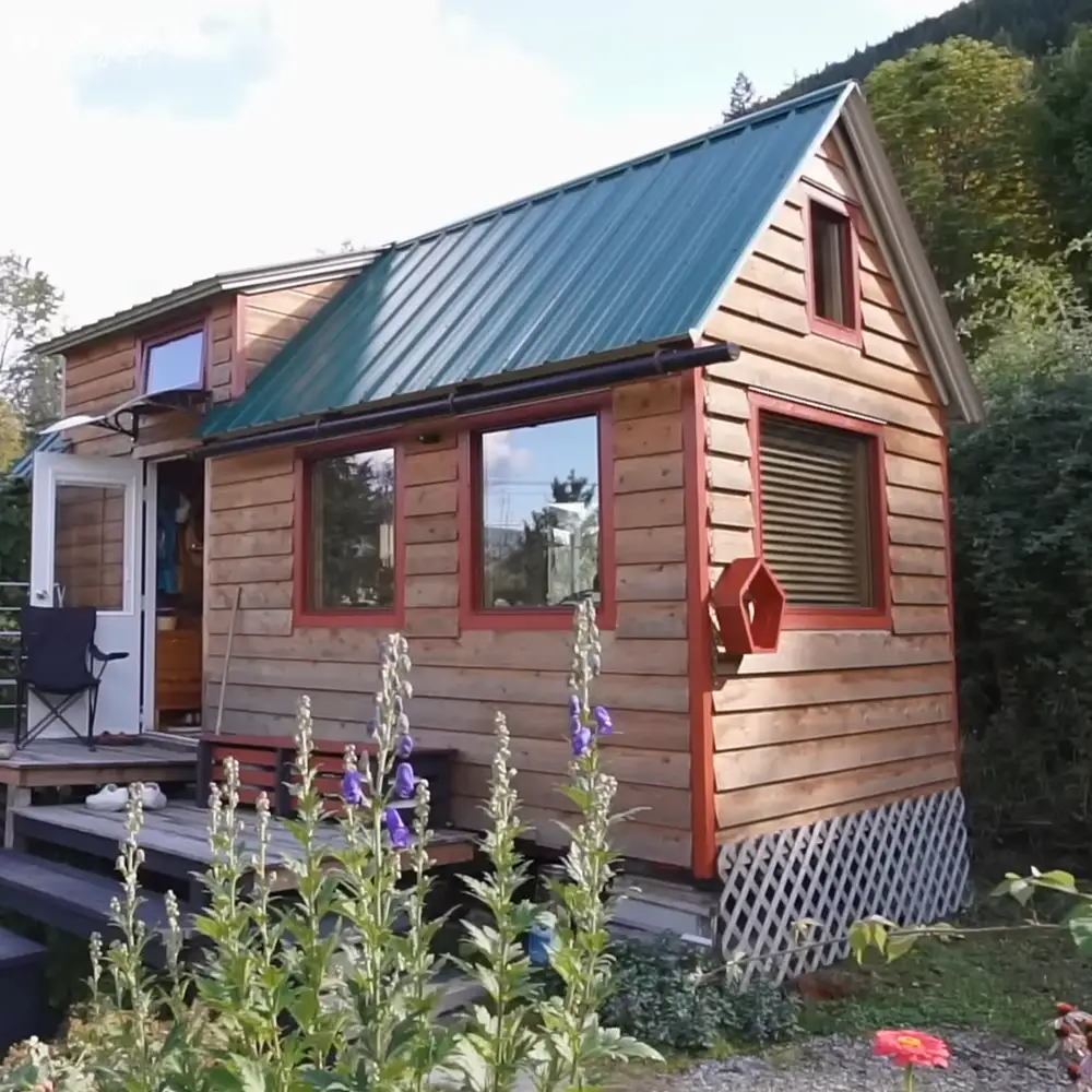 A small wooden cabin with red trim, nestled in a rural setting surrounded by greenery and blooming purple flowers.