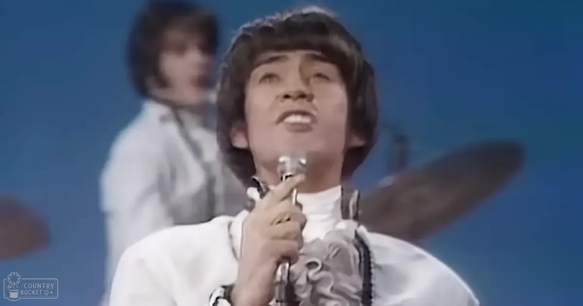 A young adult male singer wearing a ruffled white shirt performs on stage with a child to young adult (possibly teen) male, dressed in a fitted white shirt and tie, against a blue backdrop, suggesting an indoor performance venue from the 1960s or early 1970s.
