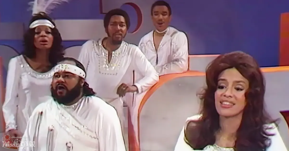 An indoor performance scene with adults wearing white outfits on a stage set with orange and blue elements, suggesting the 1970s.