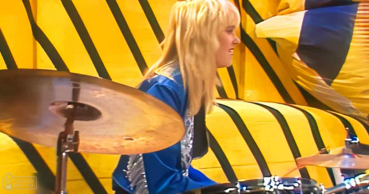 A young adult female playing drums indoors, wearing a blue uniform with silver reflective accents against a yellow wall with black diagonal stripes under bright indoor lighting.