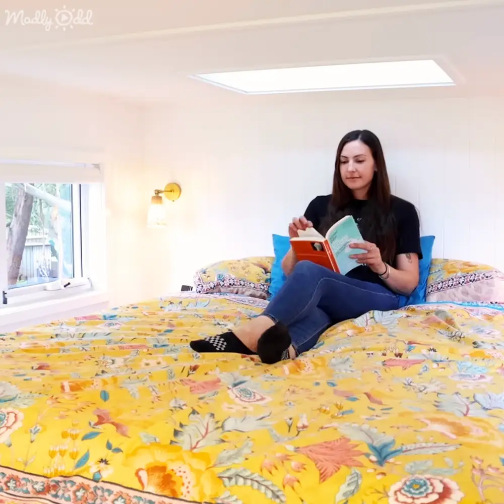 A young adult female reading in her bedroom with yellow floral-patterned bedding, blue pillows, and natural light streaming through a window showing trees and a fence.