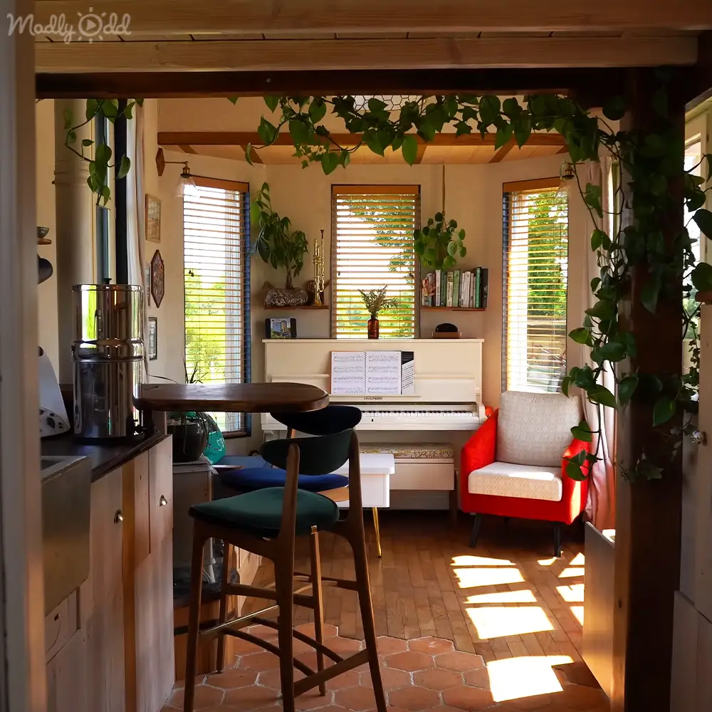 A home office or living area with a white piano, red armchair, bookshelves filled with books, plants, wooden beams, and terracotta tiled floor. The setting is illuminated by natural light streaming through horizontal blinds.