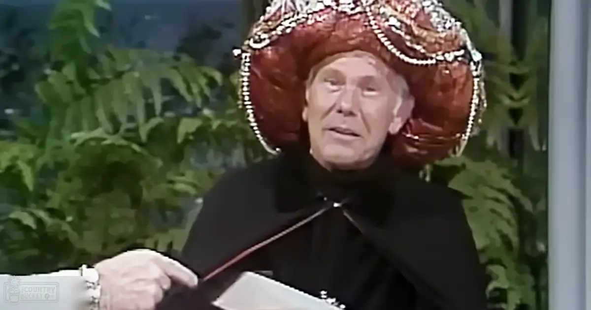 Older adult male wearing a large ornate red hat with intricate beadwork and black garment in an indoor setting with green foliage, possibly for a theatrical performance or talk show.