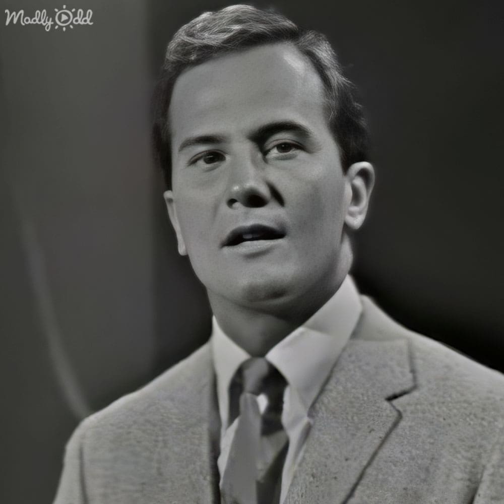 A middle-aged man in a light-colored suit jacket and white dress shirt stands against a plain dark wall.
