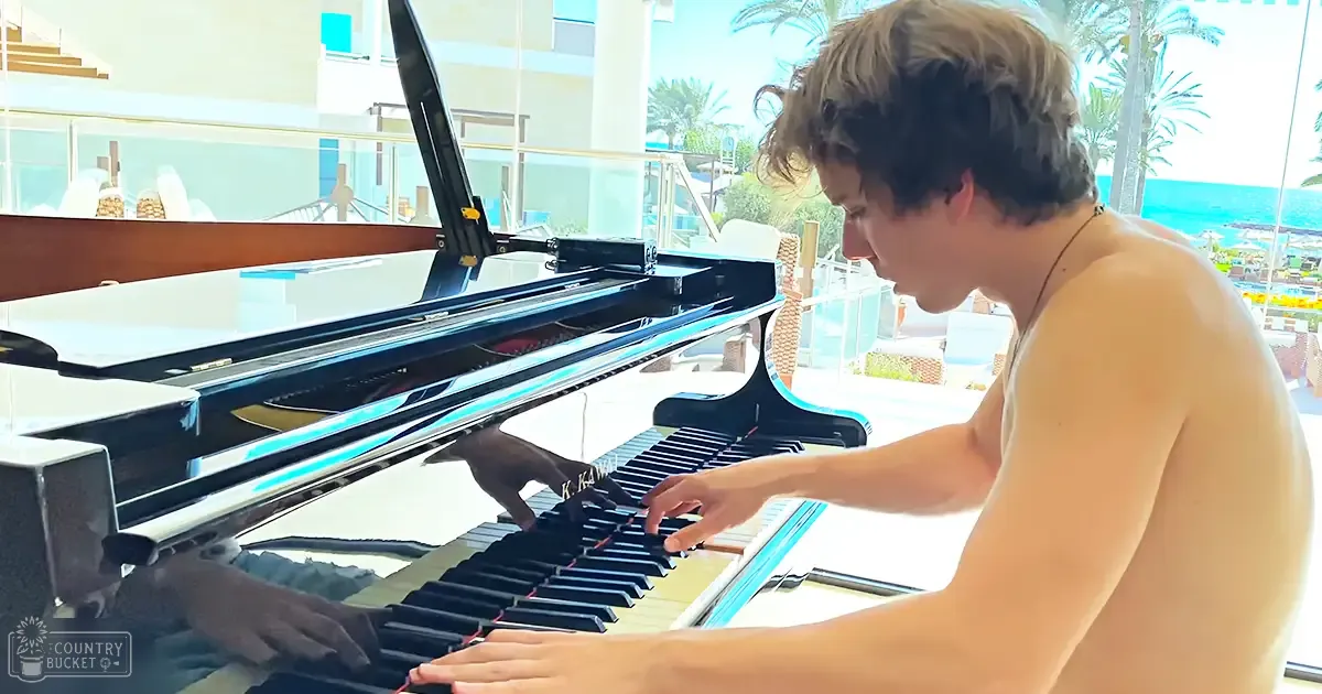 A young adult male playing a grand piano in an outdoor setting near water, surrounded by palm trees and a luxurious pool area.