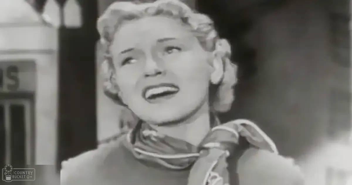 An adult female in an outdoor setting wearing a high-collared garment with a scarf or shawl draped around her neck, possibly from the mid-20th century, surrounded by an urban environment with natural daytime lighting and black-and-white tones.
