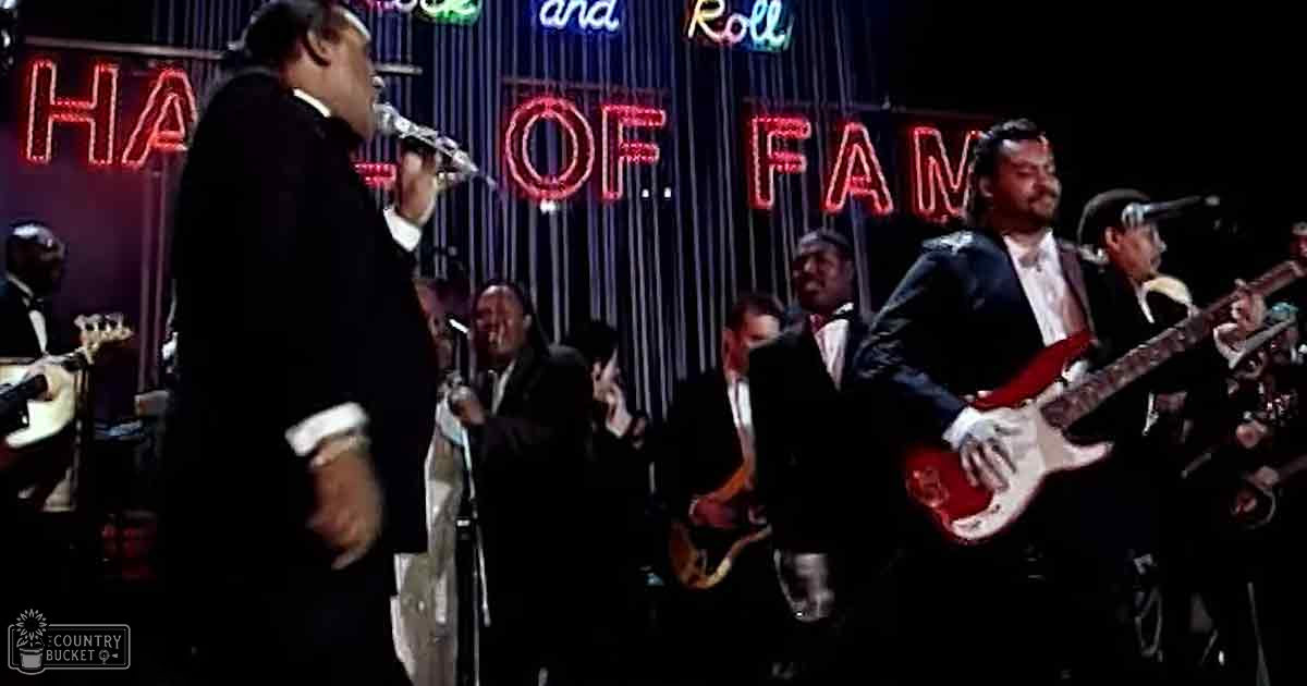 An adult male singer in a dark suit with white shirt and black tie, performing into a microphone at the Rock and Roll Hall of Fame event.